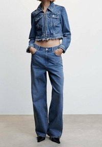 Henkilö, joka on pukeutunut korkeavyötäröisiin leveälanteisiin sinisiin farkkuihin ja lyhyeen farkkutakkiin, jossa on revitty helma, kädet taskuissa seisahtuu harmaalla lattialla.