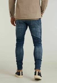Jean slim en denim bleu foncé avec une texture délavée, design à cinq poches et coutures détaillées, accompagné de baskets bleu marine et beige.