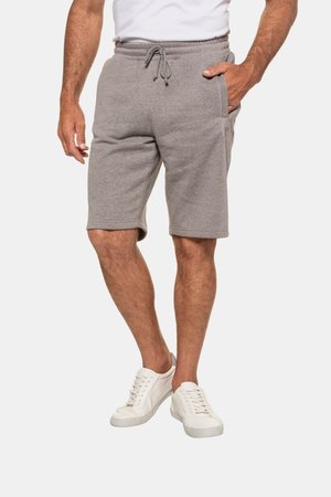 Mann trägt graue Shorts mit Kordelzug, weiße Sneakers und ein weißes Hemd, steht mit einer Hand in der Tasche vor einfarbigem Hintergrund.