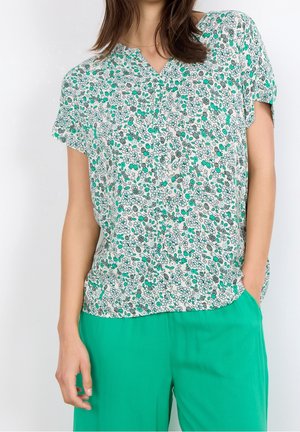 Femme portant un chemisier floral à manches courtes avec des fleurs vertes, blanches et grises, associé à un pantalon vert vif.