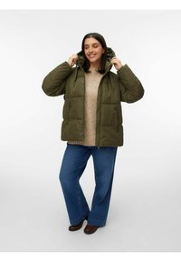 Grüne Pufferjacke mit Kapuze, vorderen Taschen und Reißverschluss, getragen über einem beigen Pullover und blauen weit geschnittenen Jeans.