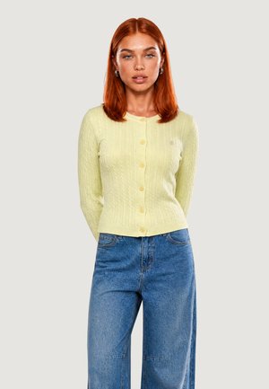 Donna con capelli rossi che indossa un cardigan giallo pallido abbottonato e jeans blu a gamba larga, in piedi davanti a uno sfondo chiaro.