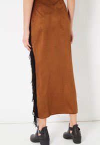 Vestido marrón de ante sintético con detalle de flecos laterales en negro. Presenta una silueta ajustada y un diseño de longitud media. Botines con tacones gruesos.