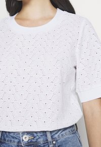 Top bianco a maniche corte con un design testurizzato e ricamato con motivi a foglia e piccoli occhielli, rifinito con un colletto rotondo.