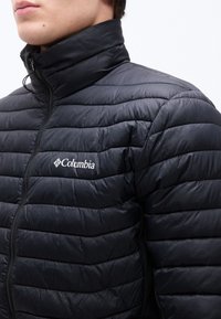 Άνδρας που φοράει μαύρο καπιτονέ μπουφάν Columbia με ψηλό γιακά, φερμουάρ κλειστό μέχρι τον λαιμό, δείχνοντας το στήθος και τους ώμους.