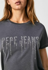 Gray cotton t-shirt featuring "PEPE JEANS" in rhinestones, with a round neckline and short sleeves. Smooth texture with subtle shine.