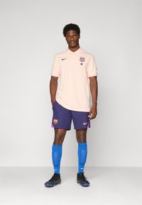 Male model standing wearing a light pink polo shirt and purple FC Barcelona shorts with blue socks and black cleats on a plain background.