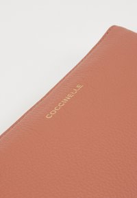 Porte-monnaie en cuir doux terracotta avec inscription dorée en relief "COCCINELLE" à l'avant, présentant une texture lisse et des bords nets.