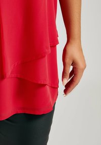 Röd skiktad blus med en flödande design, lättviktsmaterial och fållade kanter, bärs över svarta åtsittande byxor. Snygg textur och livfull färg.