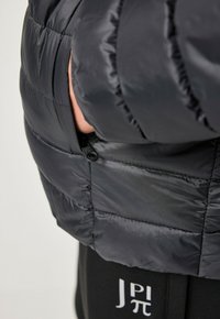 Sort puffeblæsjakke med quiltet design og sidelomme med lynlås. Glat, letvægtsmateriale, tætsiddende form med subtil glans.