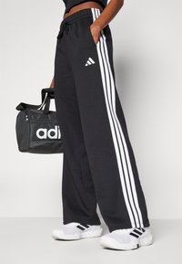 Juodos sportinės kelnės su baltomis juostelėmis, turi raištelių juosmenį ir šonines kišenes. Priedai - juoda kuprinė su "adidas" logotipu.