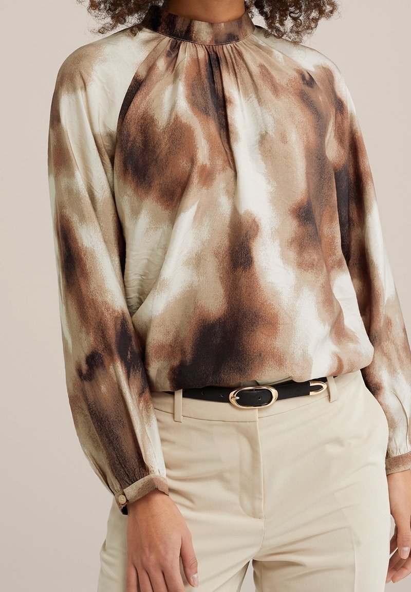 Femme portant un chemisier à manches longues et col montant avec un motif abstrait marron et crème, rentré dans un pantalon beige avec ceinture noire.
