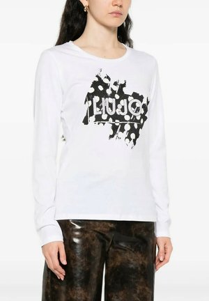 Donna che indossa una camicia bianca a maniche lunghe con motivo astratto nero e testo "LIUJO", abbinata a pantaloni marrone scuro con trama.