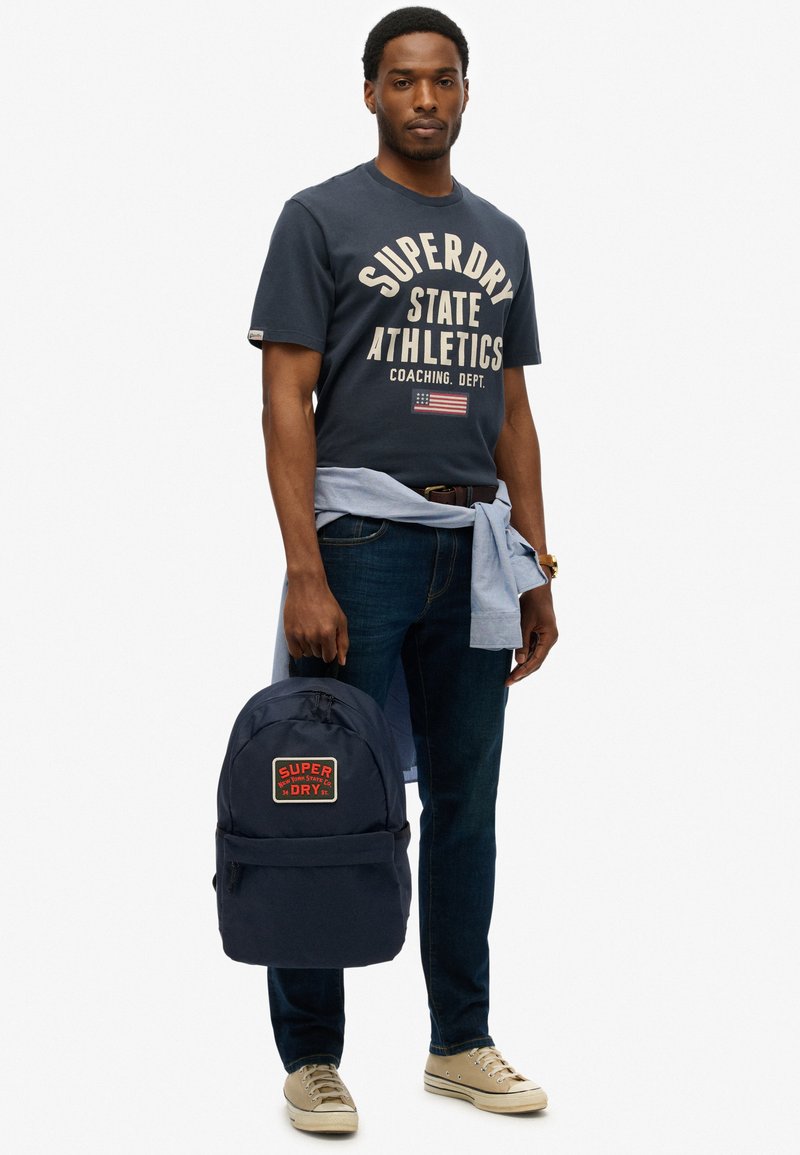 Mochila navy con parche de logotipo rojo y blanco, camiseta navy a juego, vaqueros azules y zapatos beige, sostenida por una persona en posición de pie.