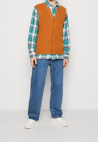 Brauner gerippter Strickwesten über einem türkis- und schwarzen karierten Hemd, kombiniert mit blauen weiter geschnittenen Jeans und weißen Sneakers. Einfaches Design, strukturierter Stoff.