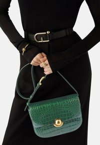 Grüne Handtasche aus krokodilpräge Leder mit einem goldenen Hardware-Verschluss und einem langen Tragegurt, gehalten von einer Person mit schwarzen langen Ärmeln und einem Gürtel.