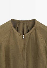 Giacca khaki con scollatura rotonda, chiusura con zip sul retro e tessuto testurizzato con orlo leggermente sfilacciato lungo il colletto.