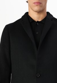 Cappotto in misto lana nera con colletto a punta, chiusura con un unico bottone e una texture sottile. Indossato sopra una polo nera con bottoni.