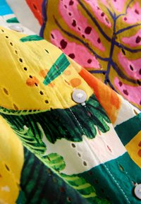 Tela colorida con patrones de hojas en amarillo, verde, rosa y naranja. A lo largo del borde, se observan botones blancos y ojales recortados.