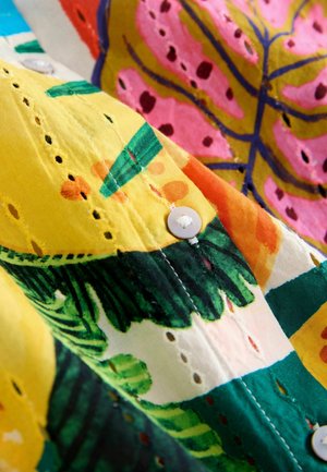 Buntes Stoffmuster mit Blattmotiven in Gelb, Grün, Pink und Orange. Am Rand sind weiße Knöpfe und Ösen-Aussparungen zu sehen.