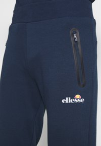 Mörkblå sweatpants tillverkade av mjukt material, med en svart ficka med dragkedja och vitt "ellesse"-logotyp med en färgglad solgrafik.