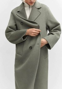 Long manteau croisé en tissu doux de couleur vert olive avec de grands revers, deux boutons et des poches latérales, porté sur un pull blanc.