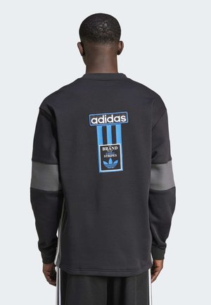 Muž nosící černou mikinu s dlouhým rukávem adidas s modrým logem a textem "The Brand with Three Stripes" na zádech, otočený zády.