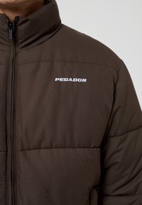 Brun puffjacka med hög krage, sidofickor med dragkedja och vit "PEGADOR"-logotyp på bröstet. Har en kviltad textur och en slank design.