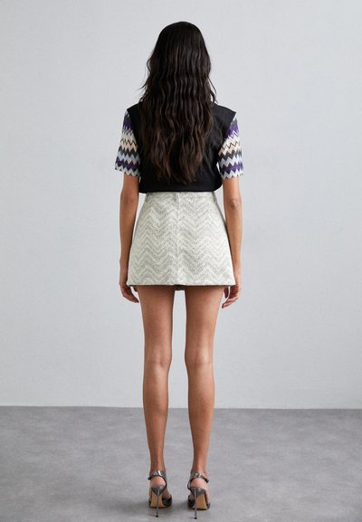 Haut sans manches noir avec des manches à motifs en zigzag, associé à une jupe blanche courte au design texturé en zigzag. Talons aiguilles visibles.