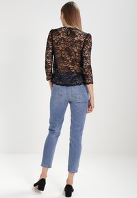 Top in pizzo nero con maniche a tre quarti, motivo floreale trasparente e orlo scallop, abbinato a jeans blu chiaro e scarpe nere con tacco.