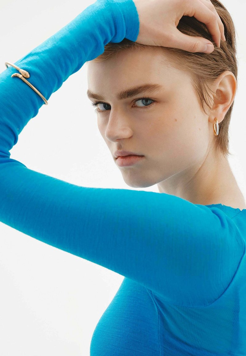 Un primer plano de un rostro con cabello corto, que lleva una blusa de manga larga de un azul brillante. Detalles notables incluyen una pulsera dorada en forma de serpiente y un arete de aro.