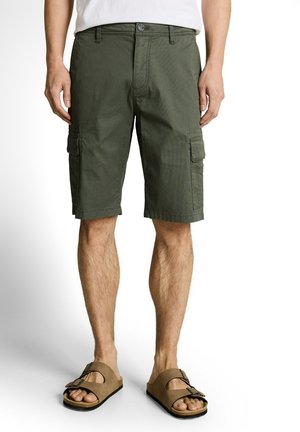 Homme portant un short cargo vert olive et des sandales à boucle beige, debout devant un fond blanc.