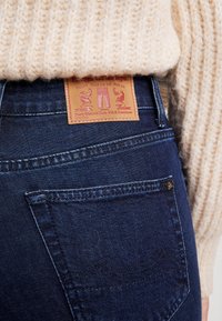 Mörkblå denimjeans med en läderlogotyp som har illustrationer. Tyget har en slät yta och en klassisk femficksdesign.