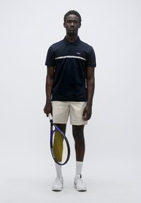 Μπλε ναυτικό πόλο με οριζόντια λευκή ρίγα, μπεζ σορτς, λευκά αθλητικά παπούτσια και κρατώντας μια κίτρινη και μαύρη ρακέτα τένις.