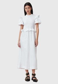 Witte jurk met een gesmokt lijfje en rimpelige peplum, met pofmouwen. De stof lijkt glad en lichtgewicht te zijn.