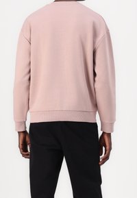 Roze sweatshirt met een ronde hals, drop shoulders, ribbelmanchetten en -zoom. Draagt met zwarte broek, met een gladde stofstructuur.