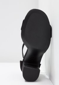 Sandale noire à talons hauts avec une semelle texturée, présentant un dessus en daim et une sangle latérale. Talon légèrement profilé pour plus de stabilité.