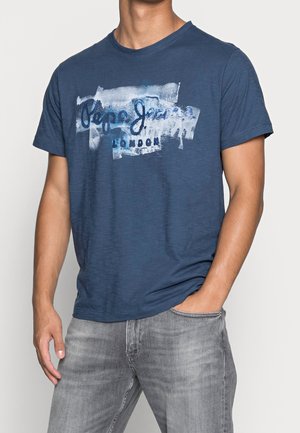 Mężczyzna w niebieskim krótkim rękawie koszulce Pepe Jeans London i szarych dżinsach, z jedną ręką w kieszeni na jednolitym tle.