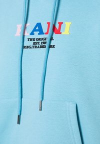 Sudadera azul claro con un bolsillo tipo canguro, cordones y un logotipo de la marca multicolor "KANI". Presenta detalles en texto negro debajo del logotipo.