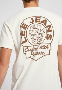 Crème-kleurig t-shirt met een grote bruine bedrukte logo op de achterkant, met de tekst "LEE JEANS Quality Denim Since 1889" en "Crafted With Purpose."