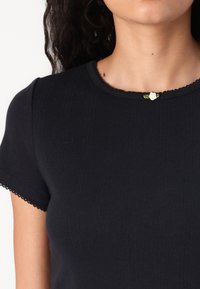 Juoda, raižyta megzta palaidinė su trumpomis rankovėmis, turinti banguotą iškirpimą ir nedidelę baltą rožę akcentą šalia apykaklės.