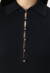 Top in maglia a coste nera con colletto, caratterizzato da un'apertura frontale decorata con sei bottoni rotondi scuri e un'apertura verticale a forma di chiave.