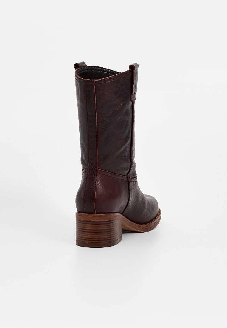 Botas de cuero marrón hasta el tobillo con un tacón grueso, que presentan un detalle de correa lateral y costuras visibles a lo largo de la parte trasera y los lados.