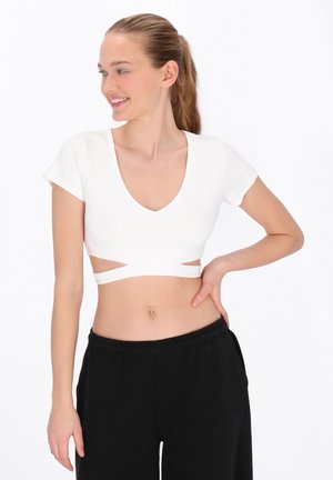 Młoda kobieta z kucykiem, ubrana w biały krótki top z wycięciami i czarne spodnie, uśmiechająca się i patrząca w lewo.