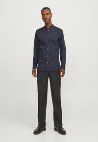 Marineblauw overhemd met knoopsluiting en een mao-kraag, lange mouwen en witte knopen. Soepele textuur, gecombineerd met donkere broeken en zwarte schoenen.