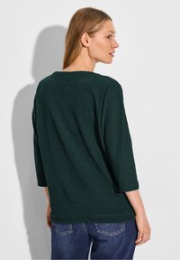 Haut texturé vert foncé avec des manches trois-quarts et un col large, associé à un jean bleu. Les coutures visibles à l'arrière mettent en valeur le design.