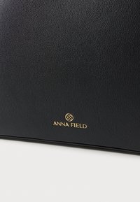 Μαύρη τσάντα από δερματίνη με υφή, με χρυσό λογότυπο "ANNA FIELD". Απλός σχεδιασμός με κομψό, ορθογώνιο σχήμα.