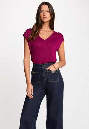 Donna che indossa una maglietta a maniche corte color magenta scintillante con scollo a V e bottoni dorati sulle spalle, abbinata a jeans a vita alta e gamba larga di colore blu scuro.