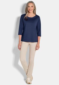 Navyblaues 3/4-Ärmeltop, runder Ausschnitt, kombiniert mit hellbeigen, eng anliegenden Hosen und cremefarbenen Turnschuhen, alles vor einem weißen Hintergrund.