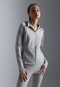 Svijetlo siva atletska zip-up dukserica s prilagođenim dizajnom, dugim rukavima i kapuljačom na vezanje, uparen s odgovarajućim uskim hlačama.